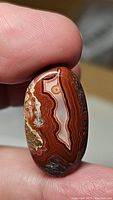 Close-up photo showing the oval cabochon shape and detailed patterning of red Crazy Lace Agate held between fingertips.
