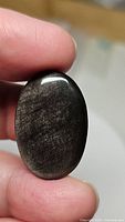Close-up photo of a polished oval cabochon silver sheen obsidian held between fingers showing the reflective silver surface and striations typical of natural obsidian stone.