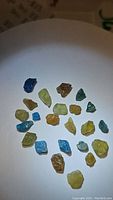 Top-down photo of 24 small rough apatite crystals in various mixed natural colors spread evenly on a white surface.