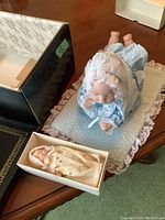 Larger blue dressed wind-up baby doll laying on blue polka dot blanket with lace edges, and smaller doll in cream outfit inside box.