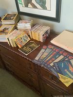 Collection of various books including Nancy Drew stacked on wooden dresser alongside magazines and protective sleeves