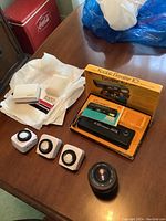 Tomkina 70-210mm lens, Kodak Ektralite 10 camera in original yellow box, three white dial timers, Kodak flash bulbs in original packaging on wooden table.