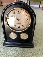 Front view of black reproduction mantel clock with round dial and two thermometer gauges below, standing on carpet.