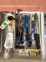 Top view of kitchen utensils including manual egg beaters, measuring cups, peelers, and knives in a white drawer organizer.