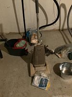 Photo showing beige upright vacuum cleaner, red and black cleaning caddy with cloths, vacuum bags, and metal bowl on floor in basement.