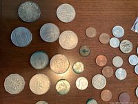 Photo shows a variety of coins laid flat on a wooden surface, including octagonal and round coins with clear and tarnished surfaces.