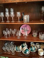 Wide view of wood cabinet shelves with assorted cut glass stemware and Royal Albert teacups on bottom shelf showing quantity and condition.