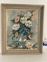 Full view of framed floral oil on board painting showing a bouquet in a white vase, three books, and one flower on the surface with a water bottle for size reference.
