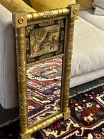 Vertical rectangular decorative mirror standing on floor beside couch, showing gold-toned frame with floral embellishments and central artwork.