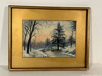Framed winter landscape painting showing snow-covered trees, small wooden bridge, and sunset sky.