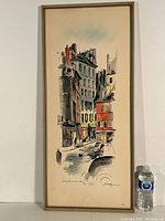 Full view of the framed watercolor painting showing the street scene with buildings and people, next to a water bottle for scale.