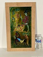 Full image of framed painted glass artwork with two stylized human figures and grapevines, next to a water bottle for scale.