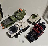 Four vintage GI Joe military vehicles on a white table including a green troop carrier, white personnel carrier, beige tank, and red/black vehicle.