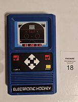 Front view of the vintage blue handheld electronic hockey game showing the screen with hockey rink graphic and LED scoreboard, control buttons including directional arrows, shot buttons, and other function keys.