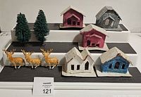 Front and angled view of 5 vintage putz houses arranged on three black steps along with 2 green bottle brush trees and 3 tan deer figurines with white spots.