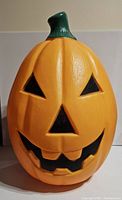 Front view of large vintage TPI pumpkin blow mold with green stem, orange body, and jack-o'-lantern face including eyes, nose, and mouth carved out to let light shine through.