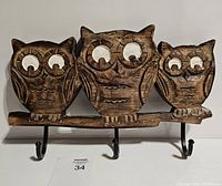 Front view of three carved wooden owls mounted on a wooden branch base with three black metal hooks below for hanging coats or other items.