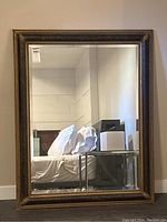 Full view of the large rectangular mirror with black and gold carved frame standing against a wall reflecting a bedroom interior.