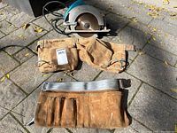 Makita circular saw with two brown leather tool belts, one lighter with gray strap and one darker with brown strap, outdoors on stone ground with dry leaves.