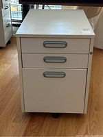 Photo of white metal filing cabinet showing front and top with all three drawers and caster wheels.