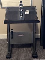 Front view of a black metal dumbbell stand with four cylindrical holders on the top surface, showing its sturdy frame and accessory clip on the side.