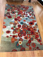 Full view of rectangular carpet with bright geometric circle pattern in red, orange, green, teal, and cream on greenish-grey background placed on wooden floor.