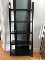 Front view of tall black leaning shelf showing five wood shelves and metal frame.