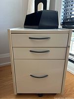 Front view of off white rolling filing cabinet with three drawers and two black plastic desk organizers on top, placed near window and wooden floor.