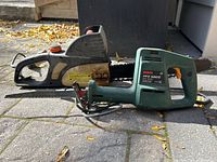 Side view of black and grey electric chainsaw and green Bosch sawzall saw on paved ground with fall leaves