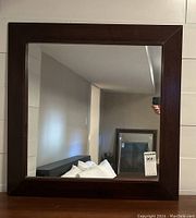 Front view of large square mirror with dark wood frame hung on wall showing beveled glass edges and mirror reflection.