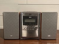 Front view of the JVC FS-S57 stereo main unit with two speakers on each side, showing control buttons and speaker covers on.