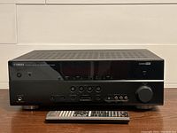 Front view of black Yamaha HTR-5063 AV Receiver with display panel showing input buttons, volume knob, headphone jack and other connectors. Remote control placed in front.