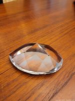 Single clear geometric cut crystal paperweight placed on wood surface showing faceted top and flat base.