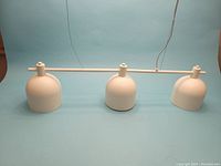 White EQ3 IYLA pendant light fixture with three bell-shaped shades displayed on a blue background, showing front view along the length of the fixture