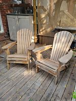 Photo showing the front and angled side of two natural wooden folding Muskoka chairs on a wooden deck, highlighting their slatted seat, back, and wide armrests with weathered wood.