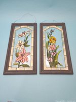 Two stained glass floral picture panels side by side in grey painted frames with a wire hanger, showing pink and white flowers.