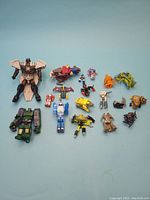 Photo showing 18 assorted vintage Transformer and robot figures laid out on blue background, including various characters from G1, G2, Beast Wars, and others. Figures vary in size, color, and condition.