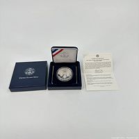 Full view showing the blue United States Mint presentation box with foam insert that holds the coin and the certificate of authenticity opened beside it.