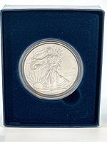 Close-up of the silver coin in protective plastic capsule inside blue velvet-lined box showing the obverse design of Liberty walking enveloped in the flag with sun rising.