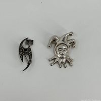 Image of two silver pins side by side on a white background showing the marcasite cat and court jester pins from the front.