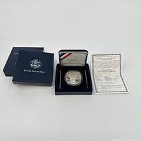 Full lot showing the silver dollar in display box, outer box, and certificate of authenticity