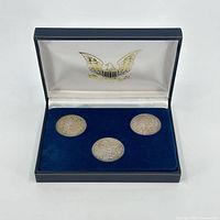 Full view of the blue display box holding three Morgan silver dollars dated 1883, 1884, and 1885. Box interior lid has a gold eagle emblem.