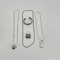 Five pieces of sterling silver jewelry including two necklaces, a cuff bracelet, and two charms laid out on white background.
