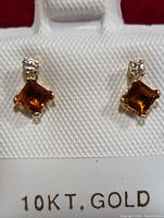 Close-up photo showing a pair of 10K gold earrings with square-cut citrine stones and small round diamonds on the post, displayed on a white textured card with '10KT. GOLD' printed below.