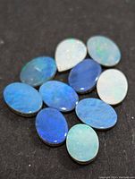 Photo showing 11 polished opal cabochons in various shades of blue and green with some play-of-color, shaped oval and teardrop, arranged on a black background.