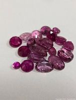 Close-up photo of multiple faceted genuine ruby gemstones in varying shapes and shades of red and pink on a white background.