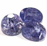 Close-up photo of three tanzanite gemstones showing round and oval cuts with visible internal inclusions and deep blue-violet color.