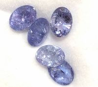 Close-up front angle of five oval faceted tanzanite gemstones in light to medium blue-violet hues on soft white fabric surface.