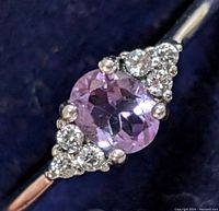 Close-up of silver amethyst ring featuring central oval faceted amethyst and surrounding clear stones on dark background.