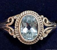 Close-up of the oval faceted blue topaz set in a twisted rope design bezel with bead and scroll accents on a silver band against a dark background.